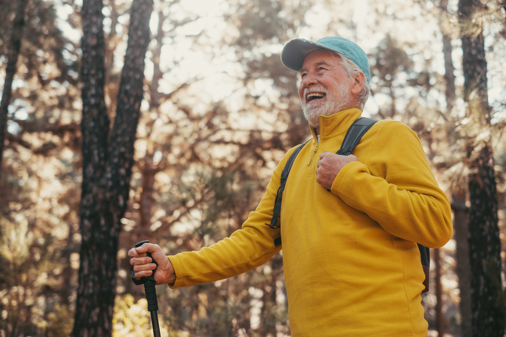 Cataract Surgery Questions Answered: Your Top FAQs_Smiling older man hiking through a sunlit forest, holding a trekking pole, free of cataracts