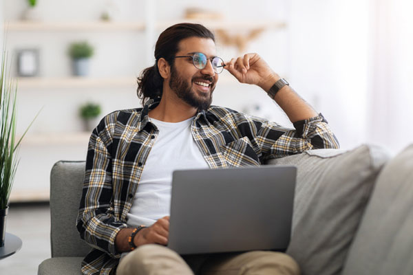 Smiling man with glasses sitting on a couch and using a laptop, representing clear, comfortable vision after astigmatism correction.