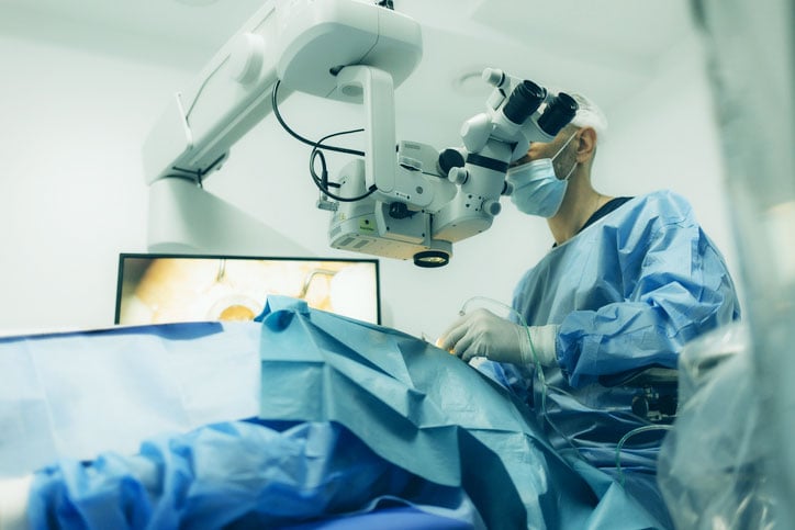 Surgical microscope and surgeon performing eye surgery in a sterile operating room, illustrating a minimally invasive glaucoma surgery (MIGS) procedure.