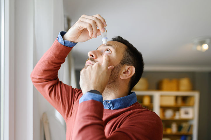 Middle-aged man applying eye drops for dry eye relief while standing near a window in soft natural light.