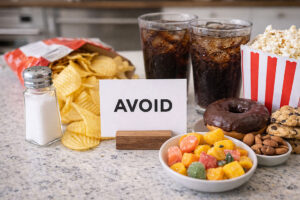 what to eat for eye health and what to avoid shown by a tabletop AVOID sign surrounded by salty chips, soda, popcorn, donuts and sugary snacks for Vantage Eye Center patients